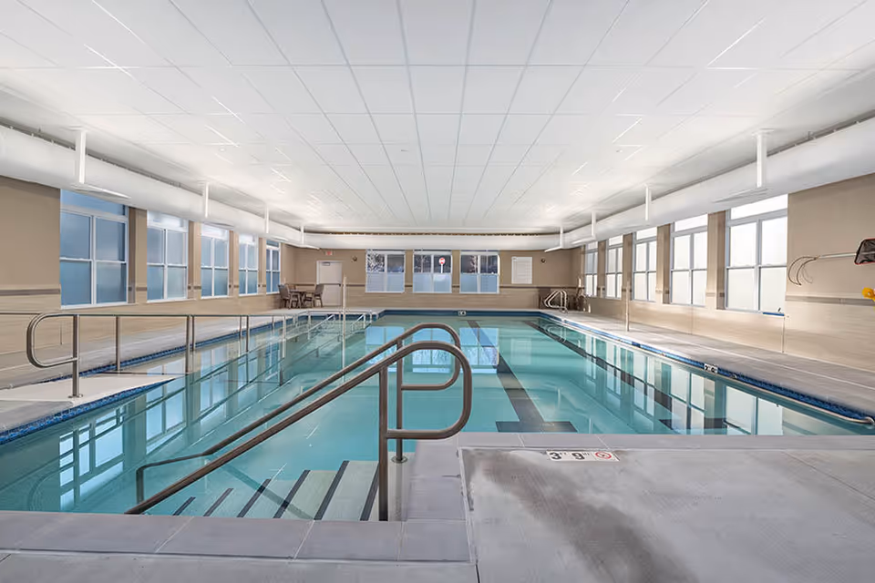 Bright indoor swimming pool with steps, handrails, and lane markings in a long windowed recreation room.