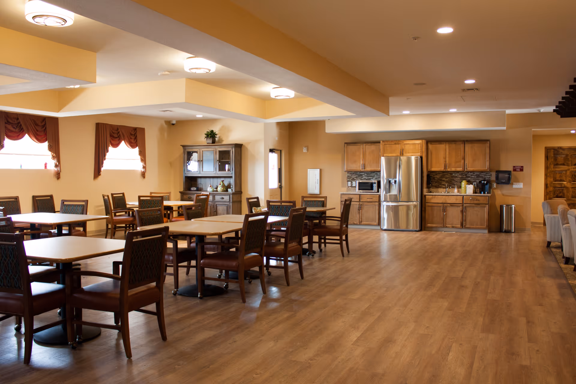 A spacious dining area in an assisted living facility featuring multiple wooden tables and chairs arranged neatly. The room has warm beige walls, wood flooring, and windows with red curtains. In the background, there is a kitchenette with wooden cabinets, a stainless steel refrigerator, a microwave, and a coffee station. The ceiling has recessed lighting and decorative light fixtures.