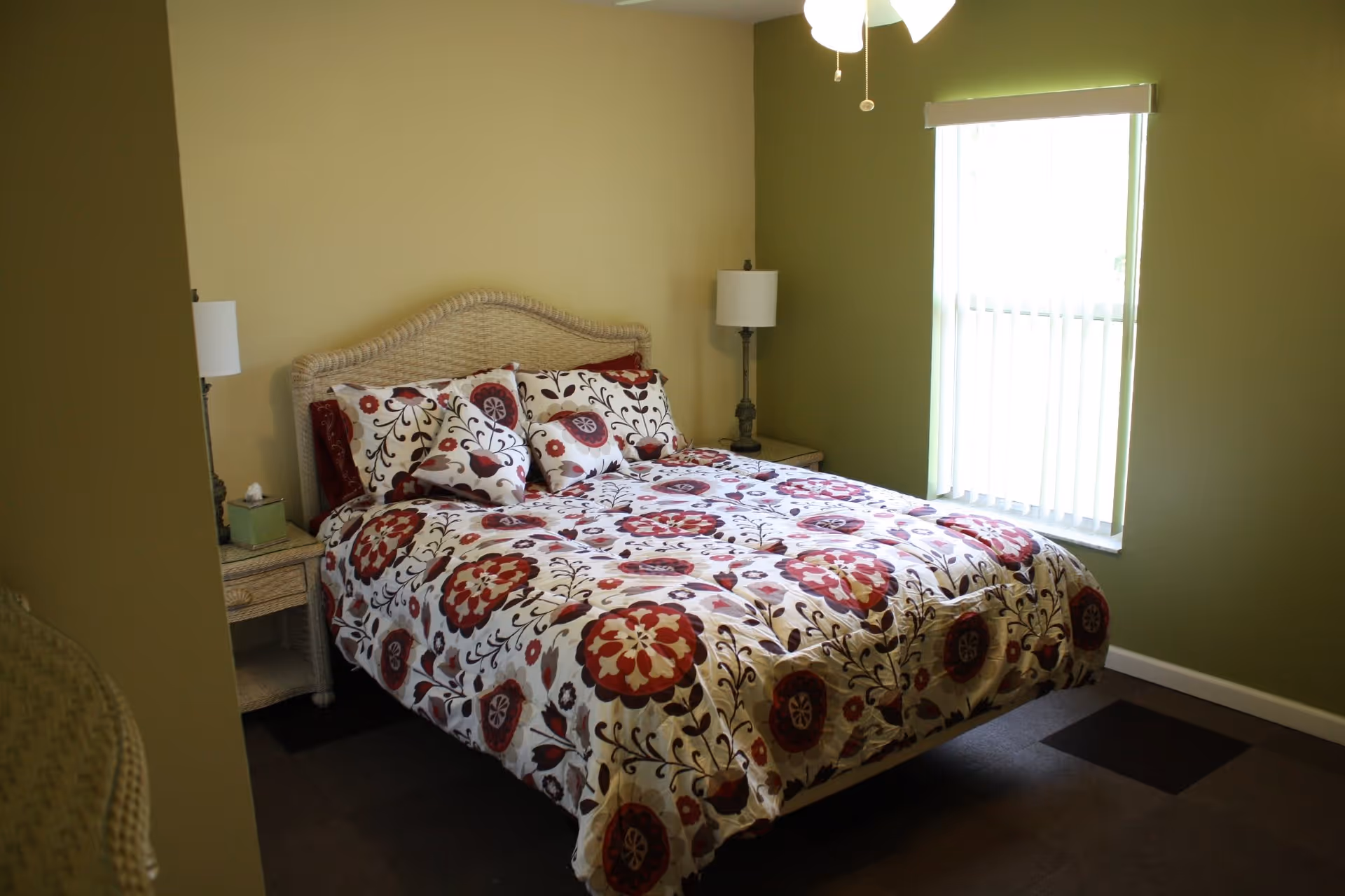 A bedroom with a bed featuring a floral patterned comforter and matching pillows. The bed has a wicker headboard and is flanked by two nightstands, each with a lamp. There is a window with vertical blinds on the right wall, and the walls are painted green.