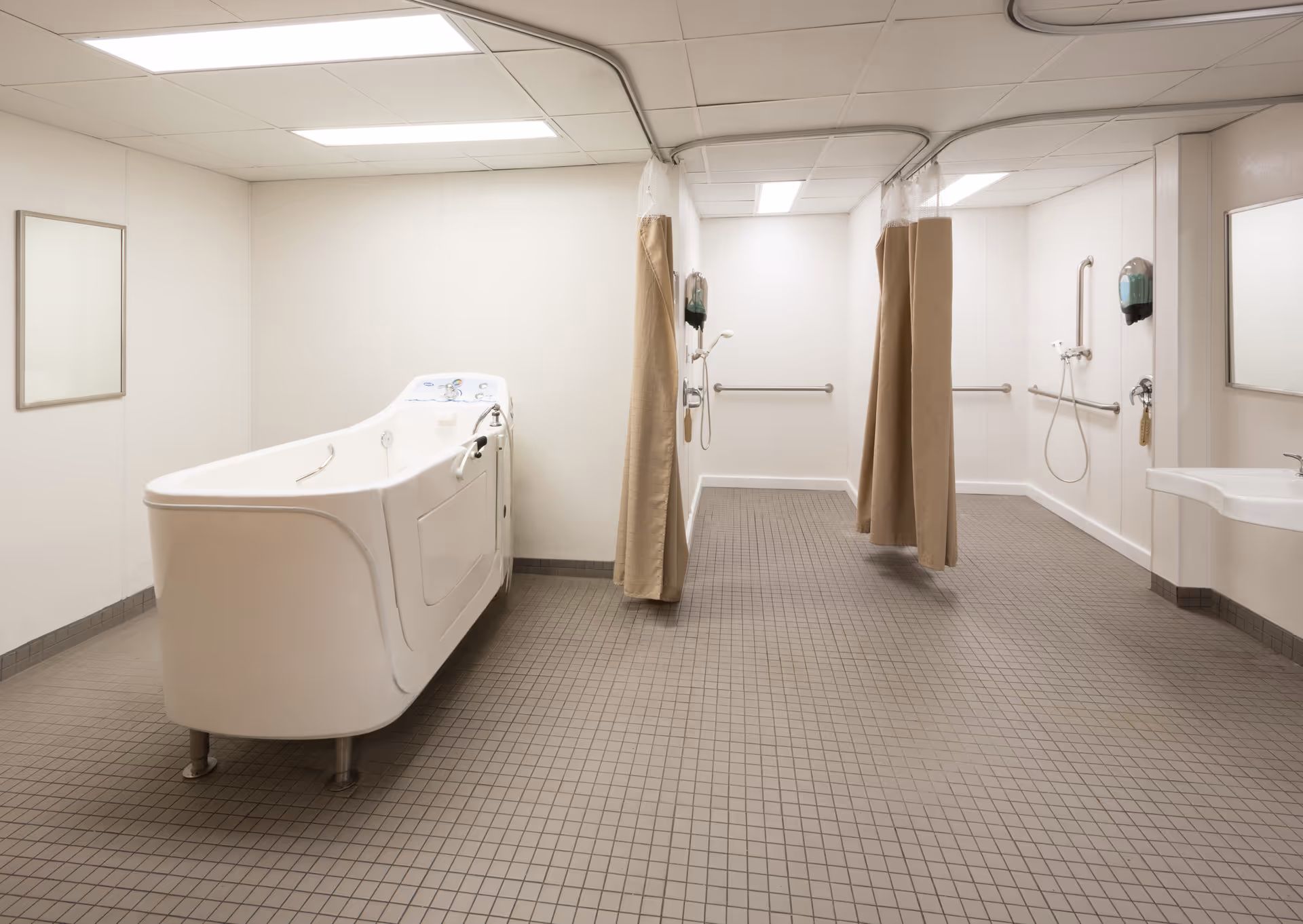 A spacious, clean bathing area in a healthcare facility featuring a large walk-in bathtub on the left, two shower stalls with beige privacy curtains in the center, and a wall-mounted sink on the right. The floor is covered with small square tiles, and the walls are plain white with grab bars installed near the showers.