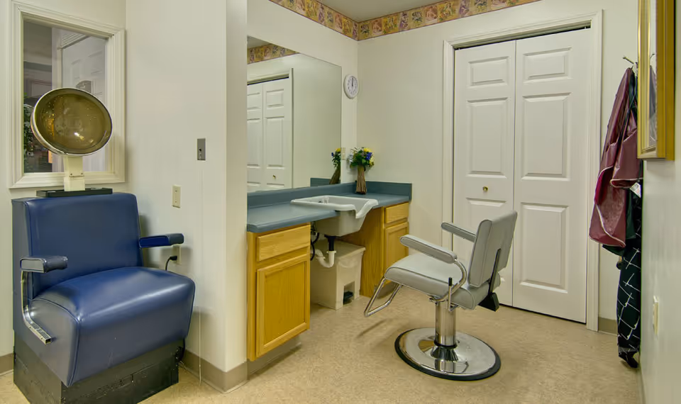 Interior of a small salon room with a blue vintage hair dryer chair on the left, a gray salon chair in front of a counter with a sink, a large mirror, and a closet with white double doors. Aprons or capes hang on hooks on the right wall.