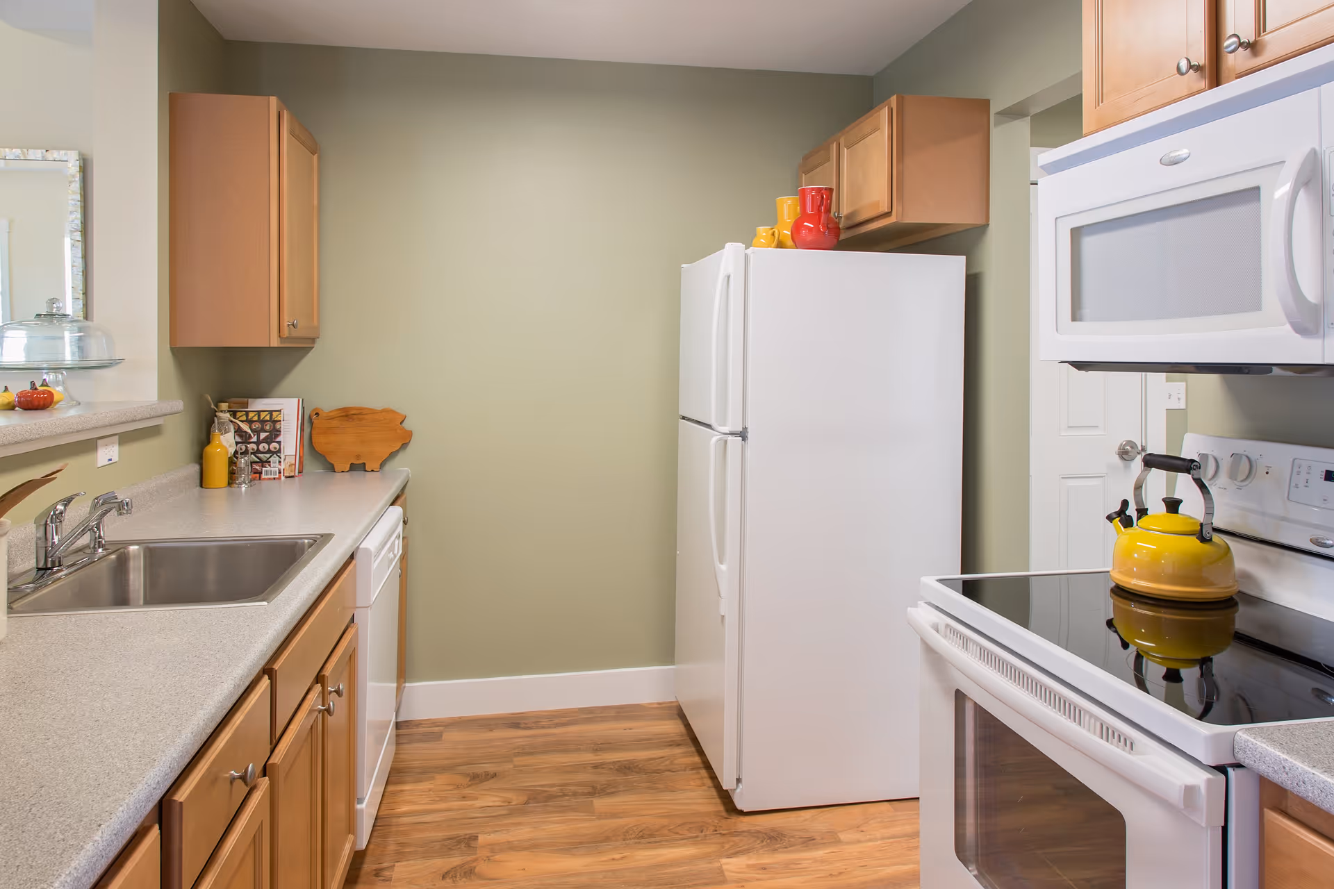 A clean and modern kitchen with light wood cabinets, a white refrigerator, white microwave, and white electric stove with a yellow kettle on top. The kitchen has a stainless steel sink, light gray countertops, and wood flooring. The walls are painted a soft green color.