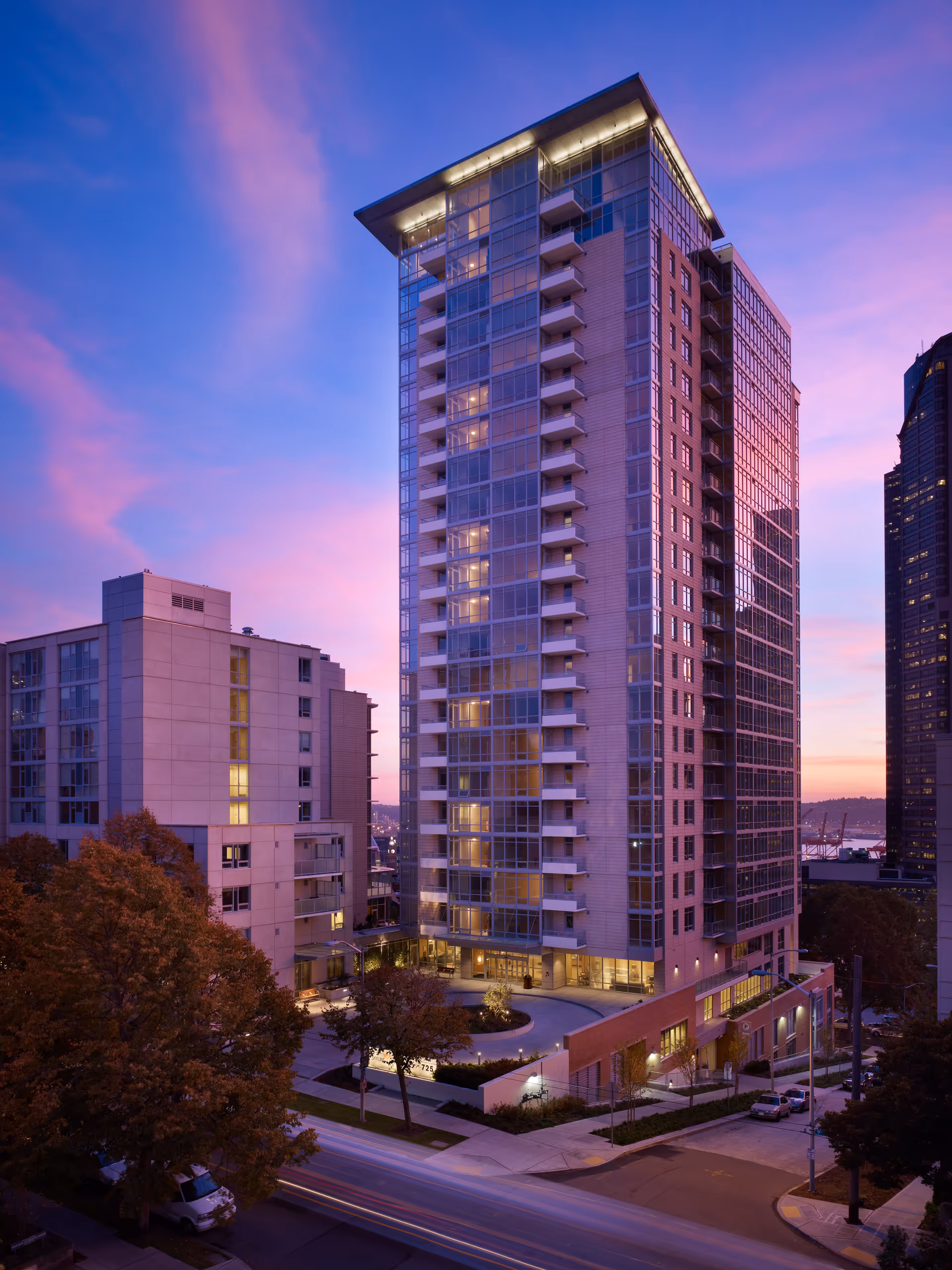Tall modern residential building at dusk with lights on inside, surrounded by other buildings and trees, under a purple and pink sky.