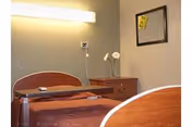 A simple senior living bedroom with a single bed featuring a brown bedspread, a wooden nightstand with a small vase holding white flowers, a wall-mounted light above the bed, and a small framed mirror on the wall.