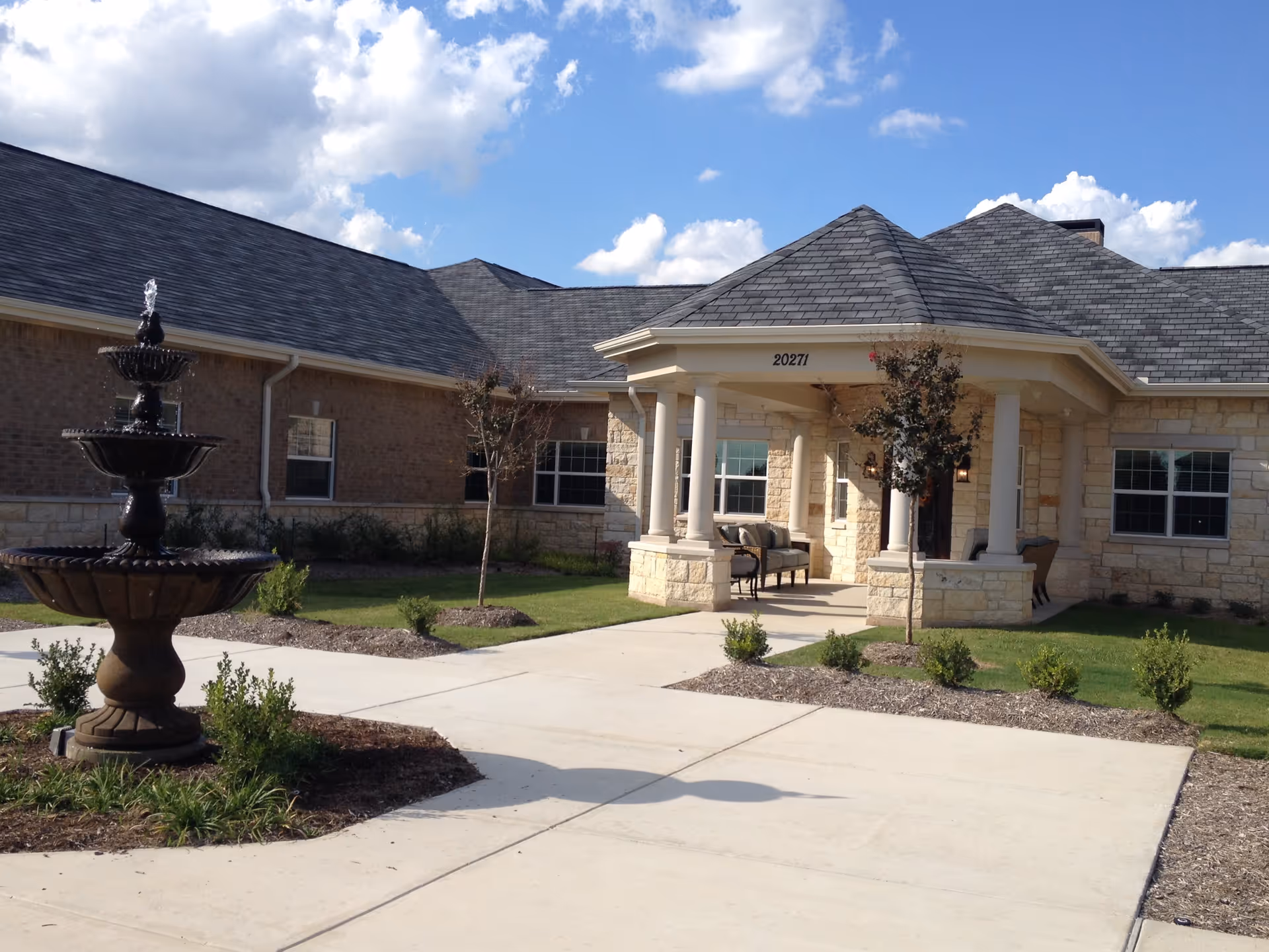 Exterior view of a single-story building with a stone facade and a covered entrance supported by columns. There is a three-tiered water fountain on the left side, surrounded by landscaped greenery and small bushes. The sky is partly cloudy with blue patches visible.