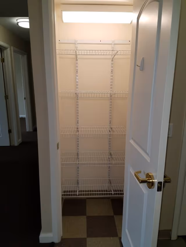 Open closet with five white wire shelves inside a small room with checkered brown and beige flooring. The closet door is white with a gold handle, and the hallway outside has dark carpet and white walls with doors visible on the left side.