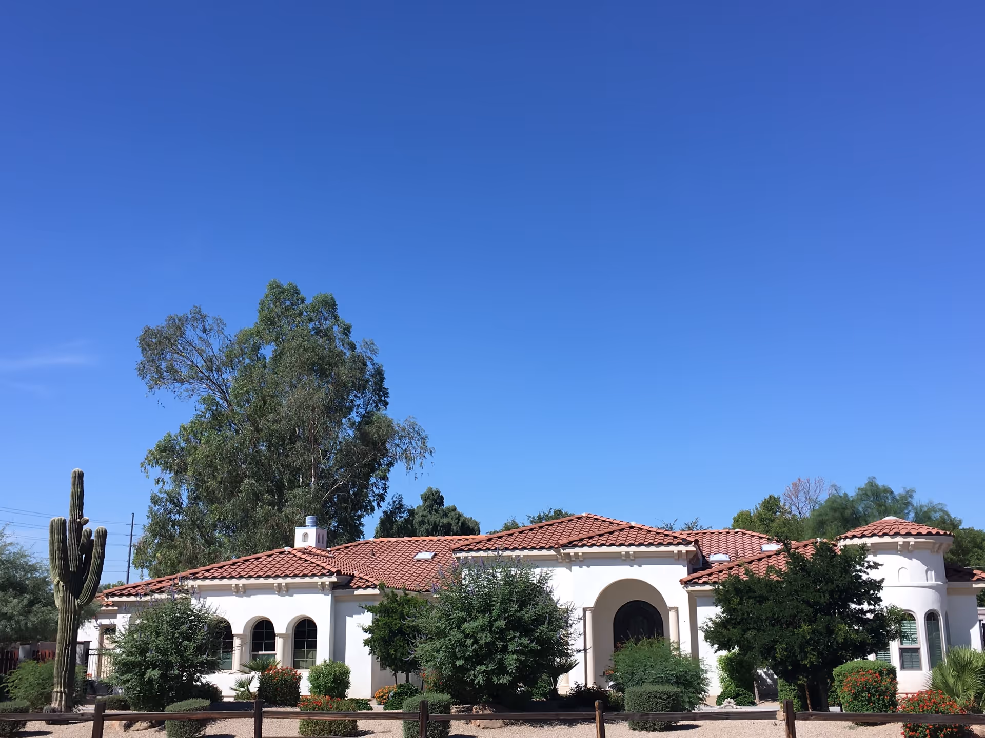 Exterior view of a single-story building with white walls and a red tile roof, surrounded by green trees and bushes under a clear blue sky.