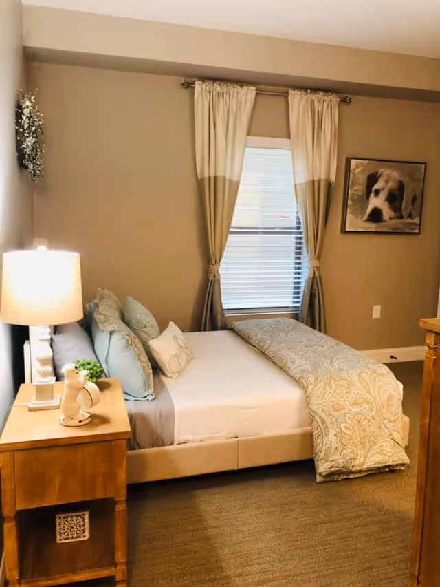 A cozy bedroom with a neatly made bed featuring light blue and white pillows and a patterned comforter. A wooden nightstand with a lamp and decorative items is beside the bed. The room has beige walls, a window with beige and white curtains, and a framed picture of a dog on the wall.