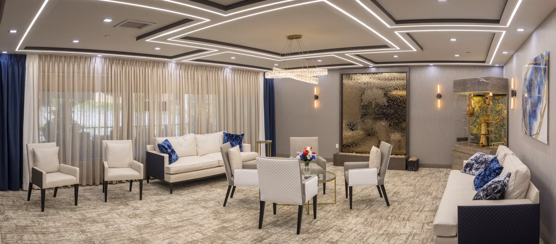 A modern and spacious living room with beige sofas and armchairs arranged around a glass coffee table with a flower vase. The room features a decorative water wall with the Dumont Center logo, blue and beige curtains, a large aquarium, and contemporary ceiling lighting.