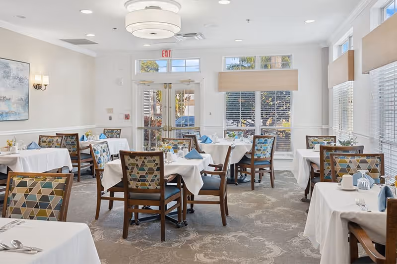 Sunlit dining room with multiple tables covered in white linens, patterned chairs, place settings, and large windows.