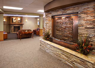 Indoor common area with a large stone wall water feature in the foreground and a seating area with a fireplace and artwork in the background.