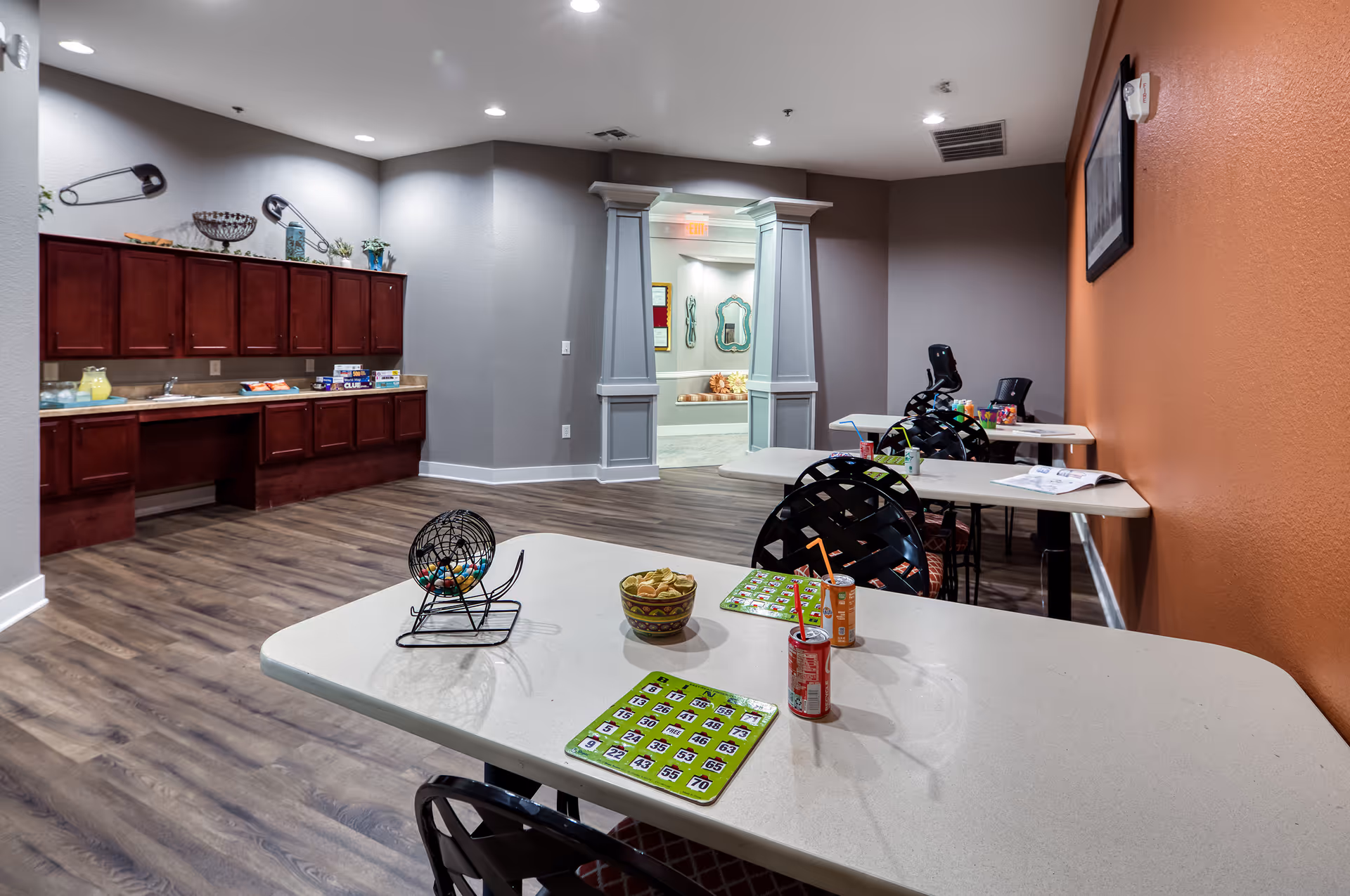 Interior common room with tables set up for bingo and snacks, a kitchenette along the left wall, and wood-look flooring.
