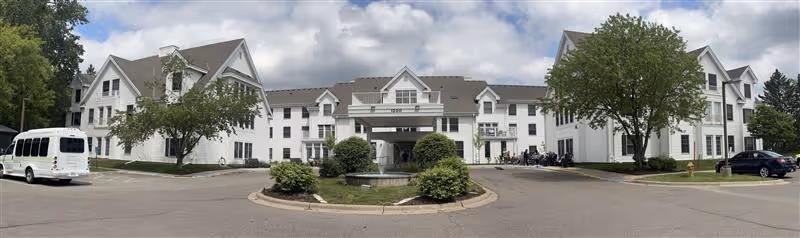 Wide panoramic view of the front exterior of a large white senior living facility building with multiple windows and gabled roofs. There is a circular driveway with a small landscaped island featuring bushes and a fountain in front of the entrance. Several vehicles, including a white van and a black car, are parked near the building. The sky is partly cloudy.
