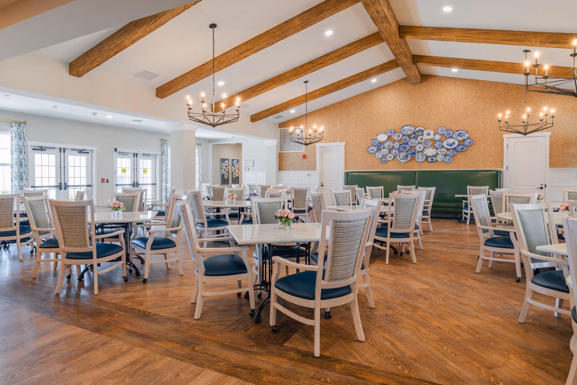 A spacious dining room with multiple square tables and chairs arranged neatly. The room features wooden floors, exposed wooden ceiling beams, and three chandeliers with candle-style lights. There is a green cushioned bench along the back wall decorated with a collection of blue and white decorative plates. Large windows and glass doors with floral curtains allow natural light to fill the room.