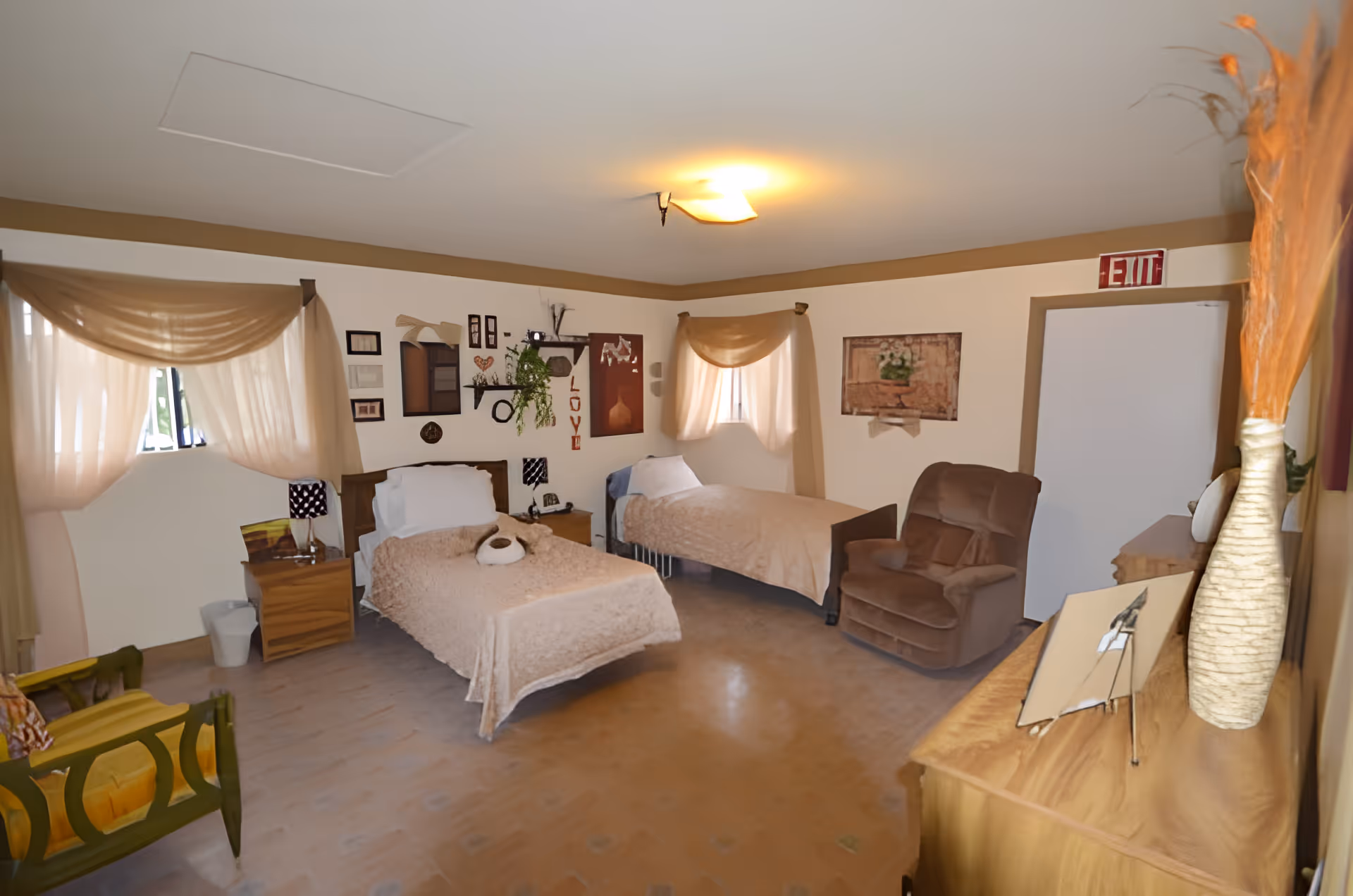 A cozy bedroom with two single beds covered in beige blankets, one with a stuffed animal on it. The room has two windows with sheer beige curtains, a brown armchair, a wooden dresser with a vase of dried plants, and wall decorations including framed pictures and shelves with plants. There is an exit sign above a door in the corner.