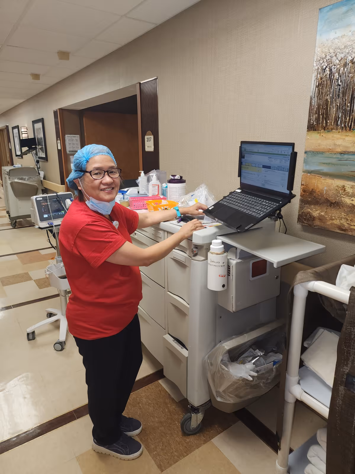 A healthcare worker wearing a red shirt, blue hair cover, glasses, and a face mask pulled down is standing next to a medical cart with a laptop on it in a hallway of a healthcare facility. The worker is smiling and appears to be working on the laptop. The hallway has beige walls, a tiled floor, and medical equipment along the walls.
