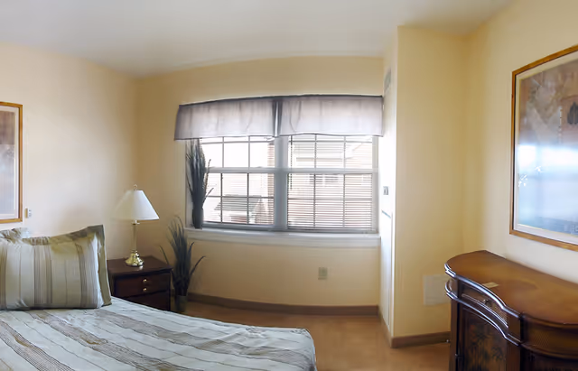 A bedroom in The Orchards Assisted Living featuring a bed with striped bedding and pillows, a wooden nightstand with a lamp, a window with blinds and a valance, a tall plant near the window, and a wooden dresser with a framed picture on the wall above it.