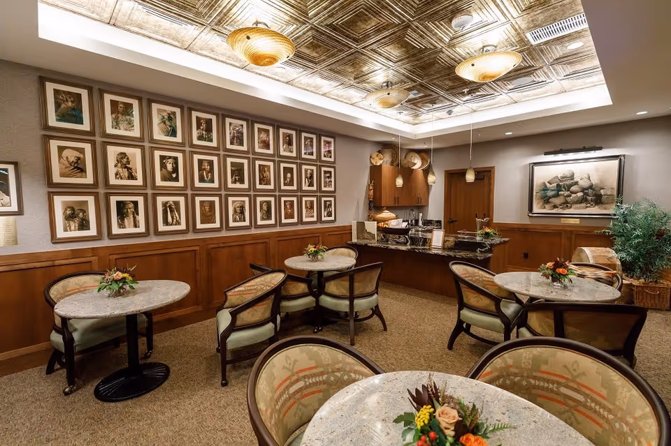 A cozy interior room with several round tables and cushioned chairs arranged for seating. Each table has a small floral centerpiece. The walls are decorated with framed portraits and a large framed artwork. The ceiling features decorative panels with warm lighting fixtures. There is a counter area with pendant lights and wooden cabinetry in the background.