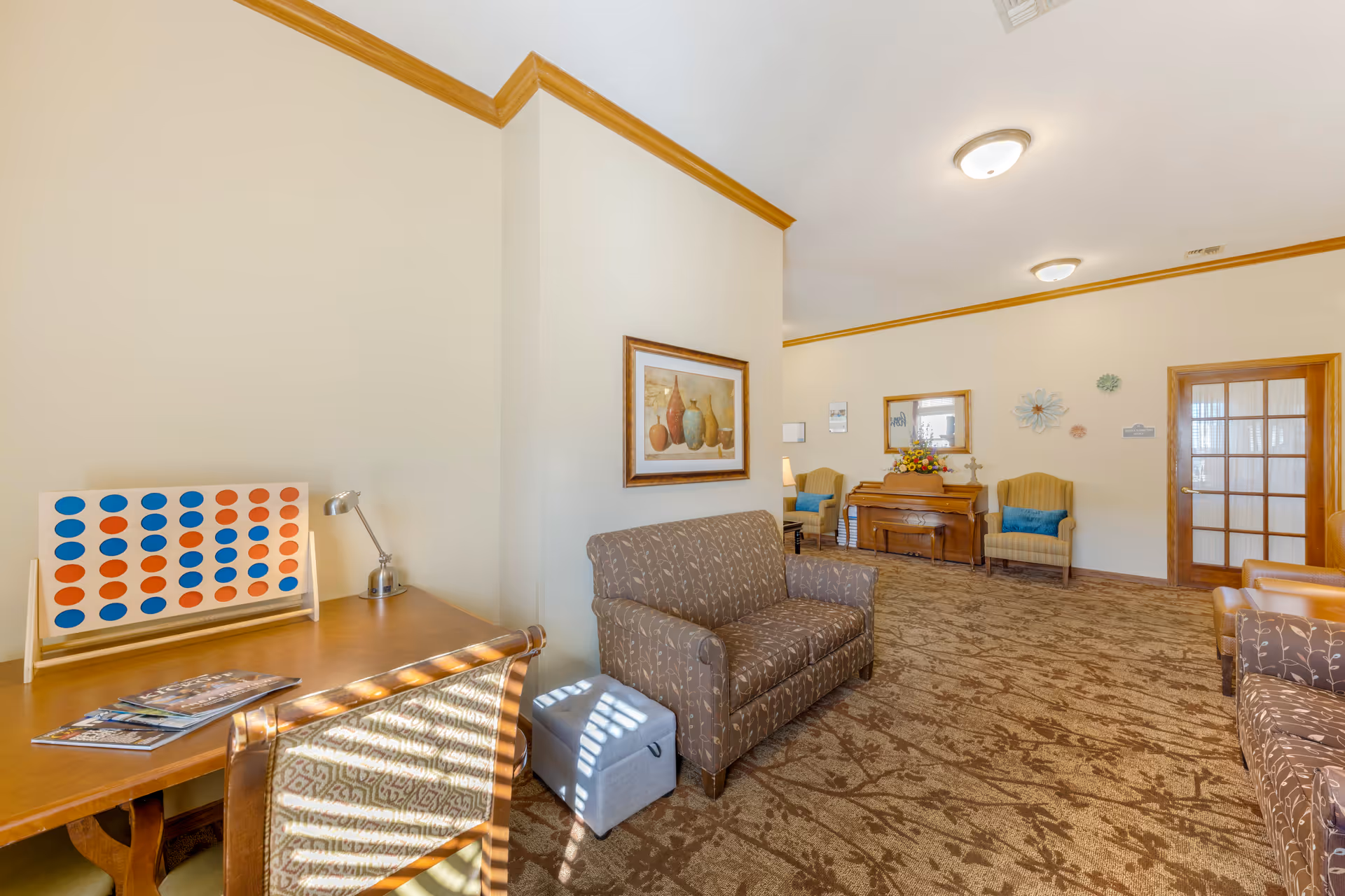 A cozy senior living facility common area with beige walls and carpet. The room features a patterned loveseat, a wooden table with a large Connect Four game, a small gray ottoman, two armchairs with blue cushions, a wooden piano with a floral arrangement on top, and wall decorations including a framed painting and decorative flowers. The space is well-lit with ceiling lights and natural light.