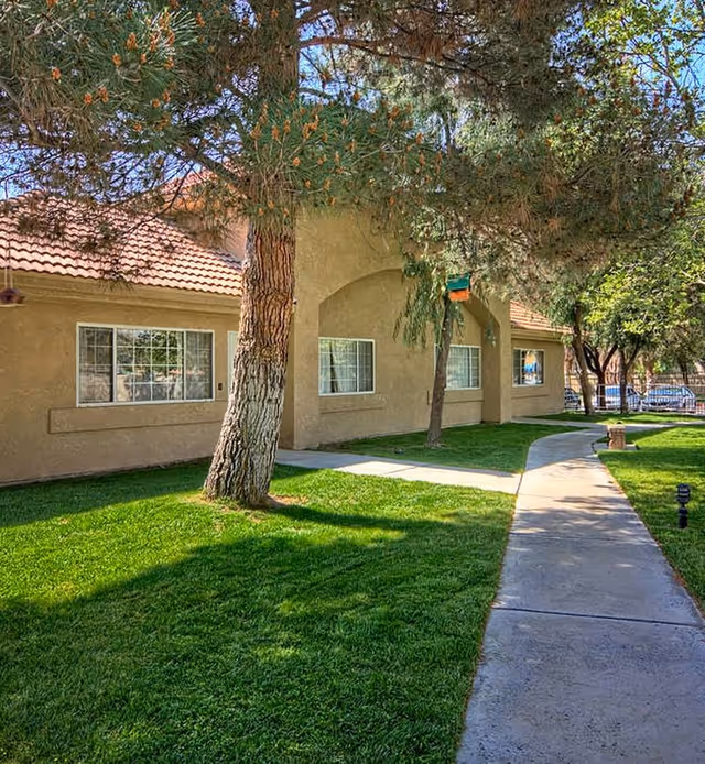 A paved walkway curves through a well-maintained grassy area with trees, leading to a beige stucco building with multiple windows and a red-tiled roof under a clear blue sky.
