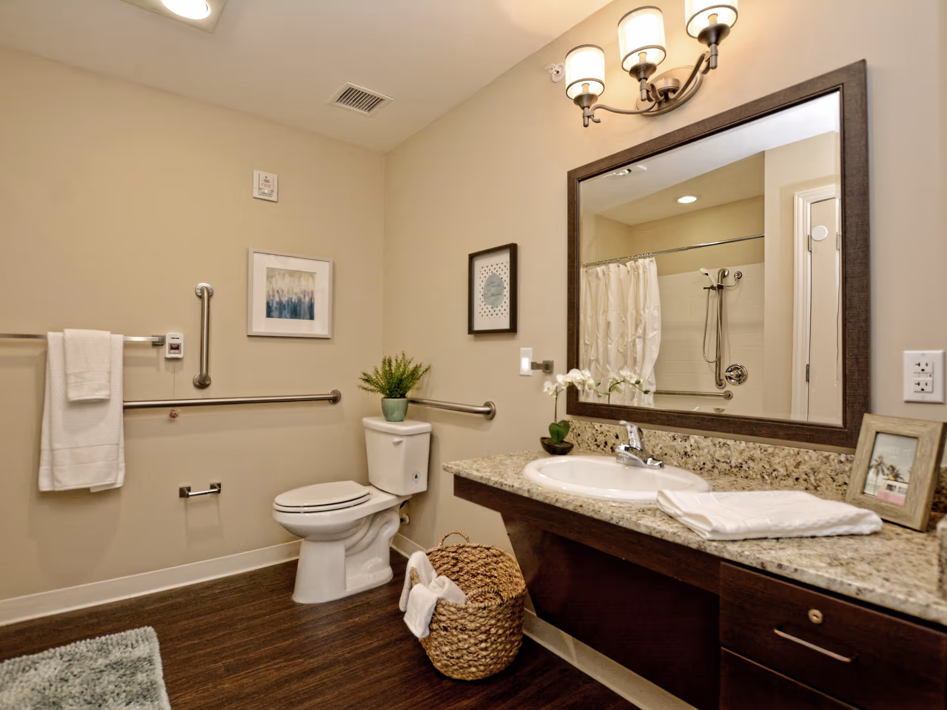 A well-lit accessible bathroom with a toilet, granite countertop sink under a large mirror, grab bars, and a shower with a curtain.