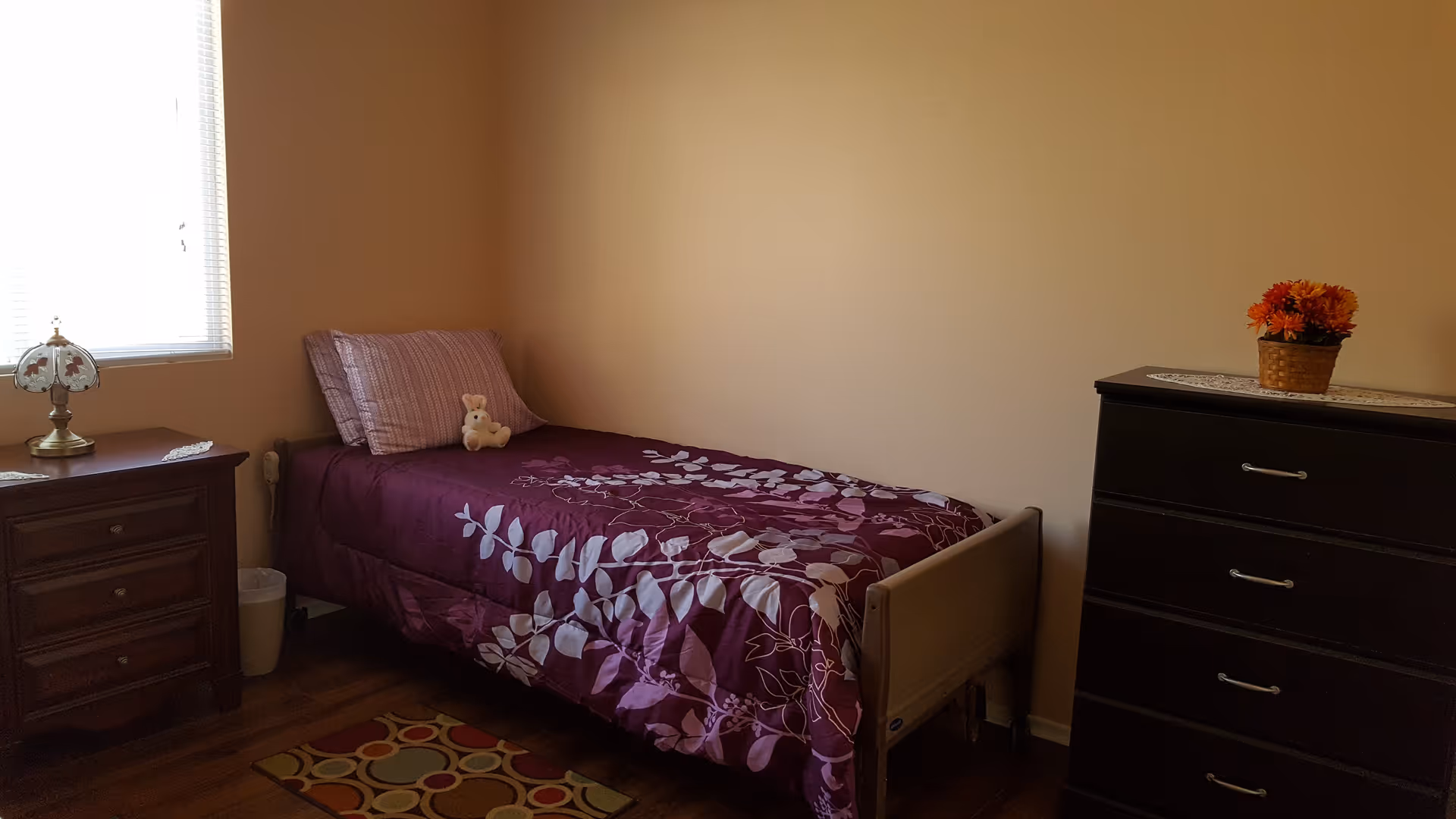 A small bedroom with a single bed covered in a purple floral bedspread and a striped pillow. A small teddy bear sits on the bed. To the left of the bed is a wooden nightstand with a decorative lamp and a small trash bin underneath. To the right is a dark wooden dresser with a basket of orange flowers on top. The room has beige walls and a window with blinds letting in natural light.