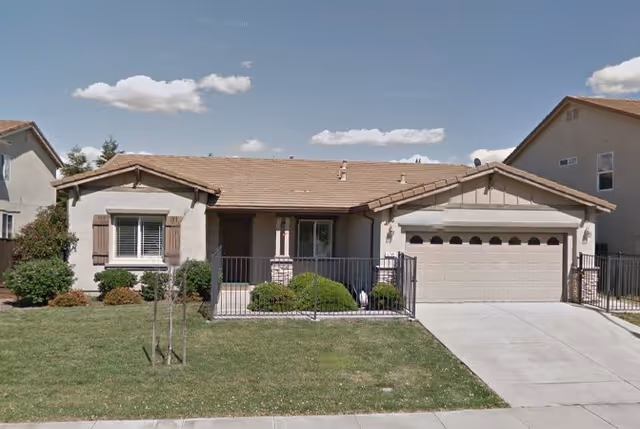 Single-story residential building with a tiled roof, a front lawn, and a driveway leading to a two-car garage. The house has a small porch area with a railing and some bushes in front.