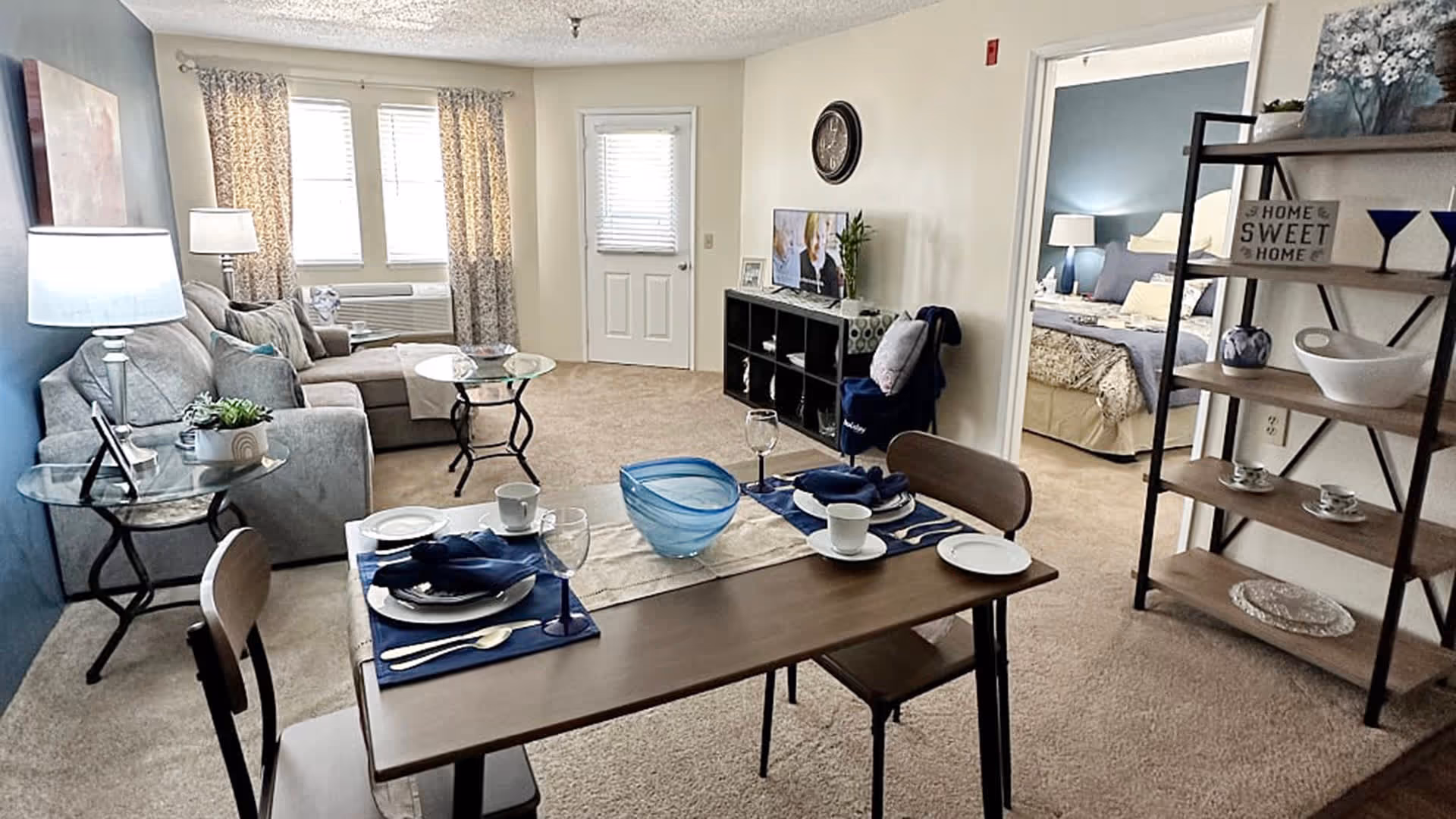 A cozy living and dining area in an apartment with a wooden dining table set for two, a gray sectional sofa, glass side tables with lamps, a TV on a black cubed shelf, and a view into a bedroom with a bed and nightstand. The room has beige carpet and light-colored walls with a blue accent wall.