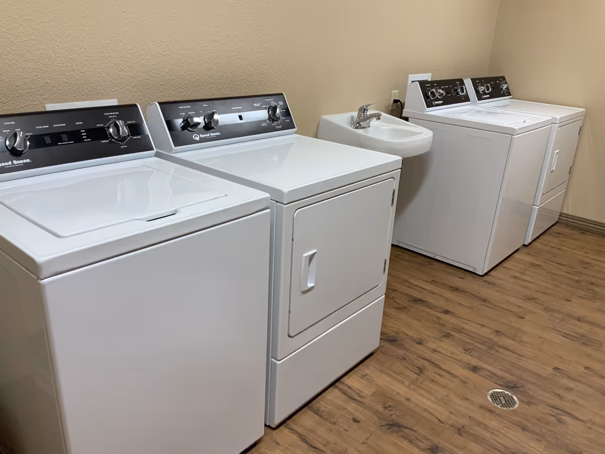 Laundry room with two washing machines and two dryers lined up against a beige wall, with a small white sink in between the machines. The floor is wood-patterned and there is a drain on the floor.