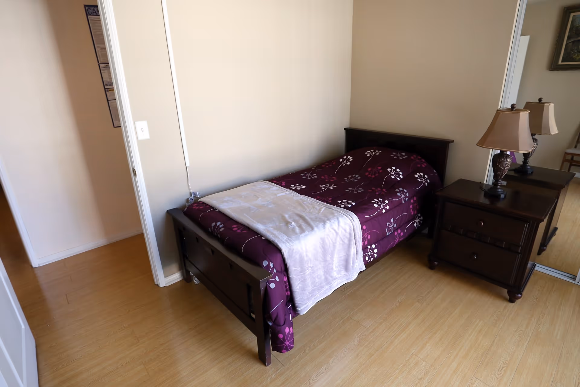 A small bedroom with a single bed covered in a purple floral bedspread and a light-colored blanket folded at the foot. Next to the bed is a dark wooden nightstand with two drawers and a decorative lamp on top. The room has light-colored walls and wooden flooring. A mirrored closet door is visible on the right side.