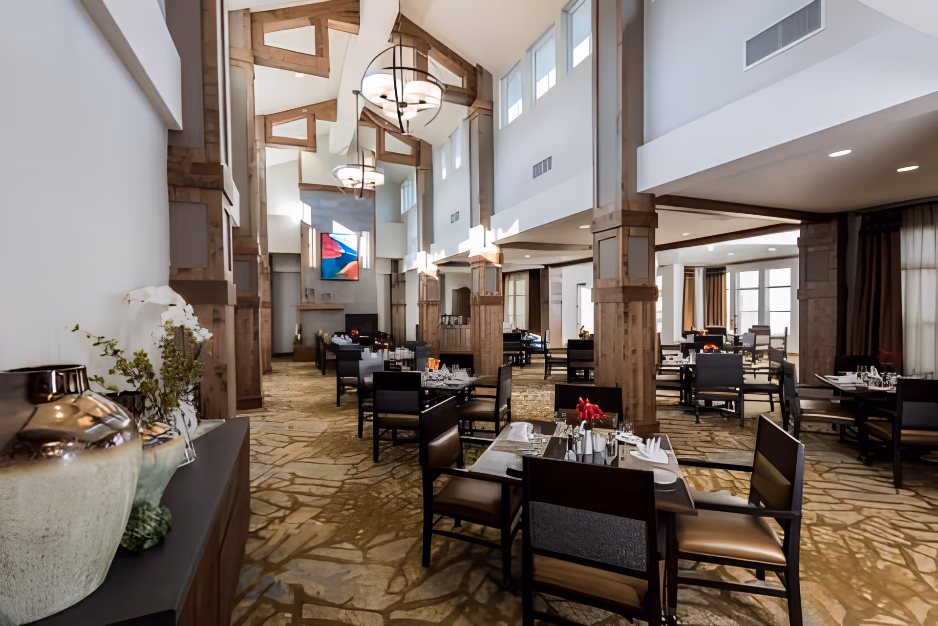 Large, elegant dining room with high vaulted ceilings, wooden columns, chandeliers and neatly set tables.