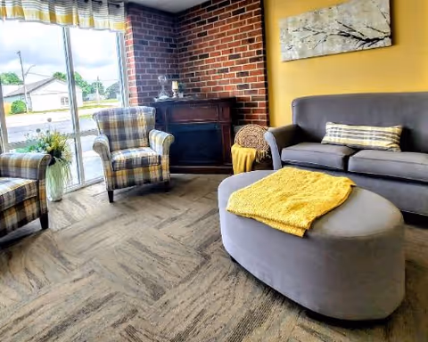 A cozy living room area with a gray sofa, a gray ottoman with a yellow throw blanket, two plaid armchairs, a small table with a flower vase, and a brick accent wall. Large windows with striped curtains let in natural light.