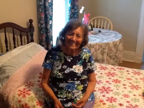 An elderly woman with gray hair wearing a floral shirt is sitting on a bed with a festive snowflake-patterned bedspread in a cozy bedroom. Behind her, there is a round table covered with a floral tablecloth and a wooden chair. A window with floral curtains is visible in the background.
