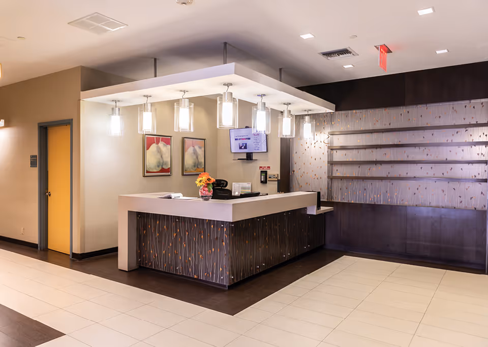 Reception area with a modern front desk featuring a dark wood pattern and a light countertop. Above the desk, there are six hanging pendant lights. Behind the desk, two framed artworks are displayed on the wall, and a small monitor is mounted. The floor has a combination of light and dark tiles, and there is a yellow door to the left.