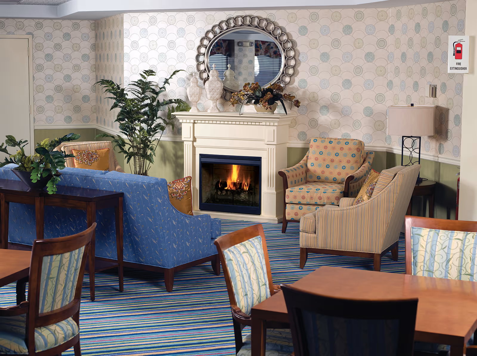 Cozy assisted living common room with a lit fireplace, upholstered chairs and sofas, dining tables, and a decorative mirror above the mantel.