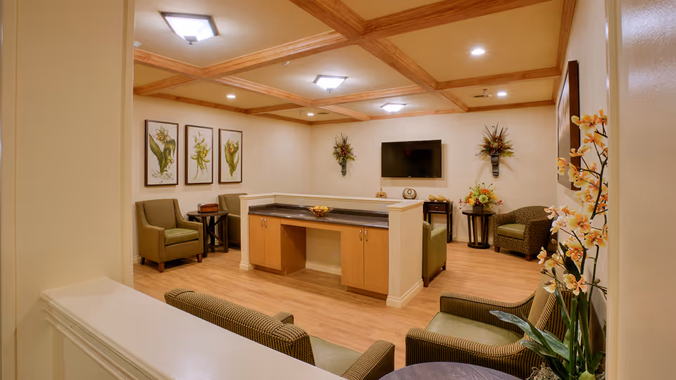 A cozy living room area in an assisted living facility with wooden flooring, beige walls, and a coffered ceiling with recessed lighting. The room features several armchairs arranged around a central wooden cabinet with a granite countertop. A flat-screen TV is mounted on the wall, flanked by decorative floral arrangements. Botanical artwork is displayed on one wall, and a vase with orange flowers is visible in the foreground.