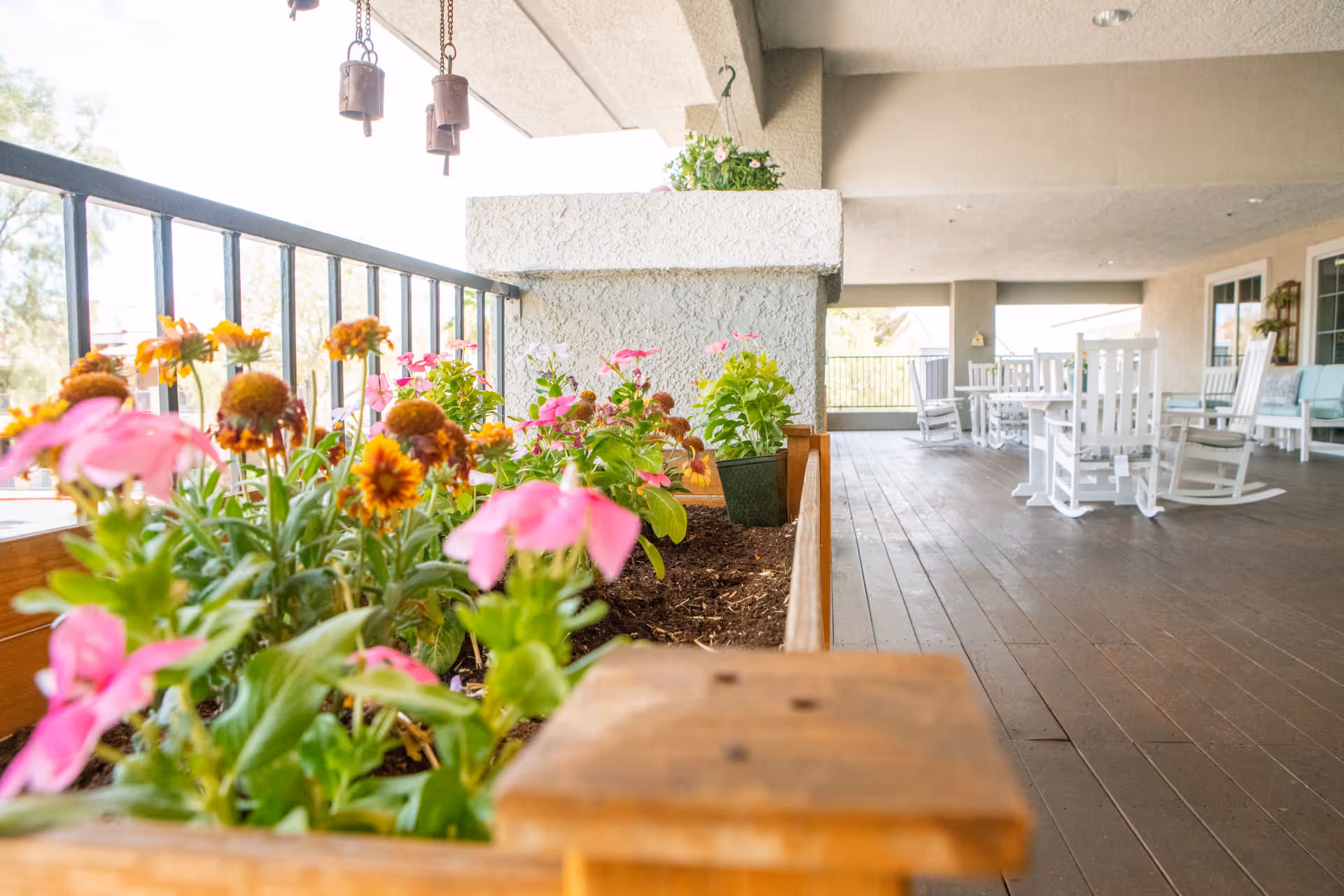 Covered outdoor patio with a wooden planter of pink and orange flowers in the foreground and white rocking chairs and tables in the background.