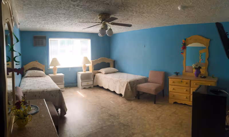 A bedroom with two single beds placed against blue walls, each bed with a light-colored headboard and floral bedspread. Between the beds is a window with blinds and two matching bedside tables with lamps. A pink upholstered chair is positioned near a yellow dresser with a mirror, decorated with a purple vase of flowers. The ceiling has a fan with lights, and the floor is a light-colored tile.