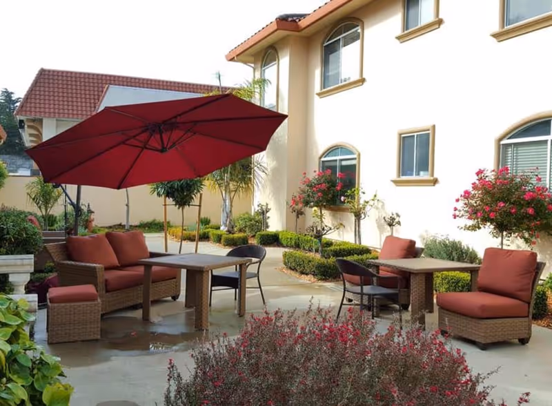 Outdoor patio area with cushioned wicker chairs and tables under a large red umbrella, surrounded by plants and flowers next to a beige building with arched windows.