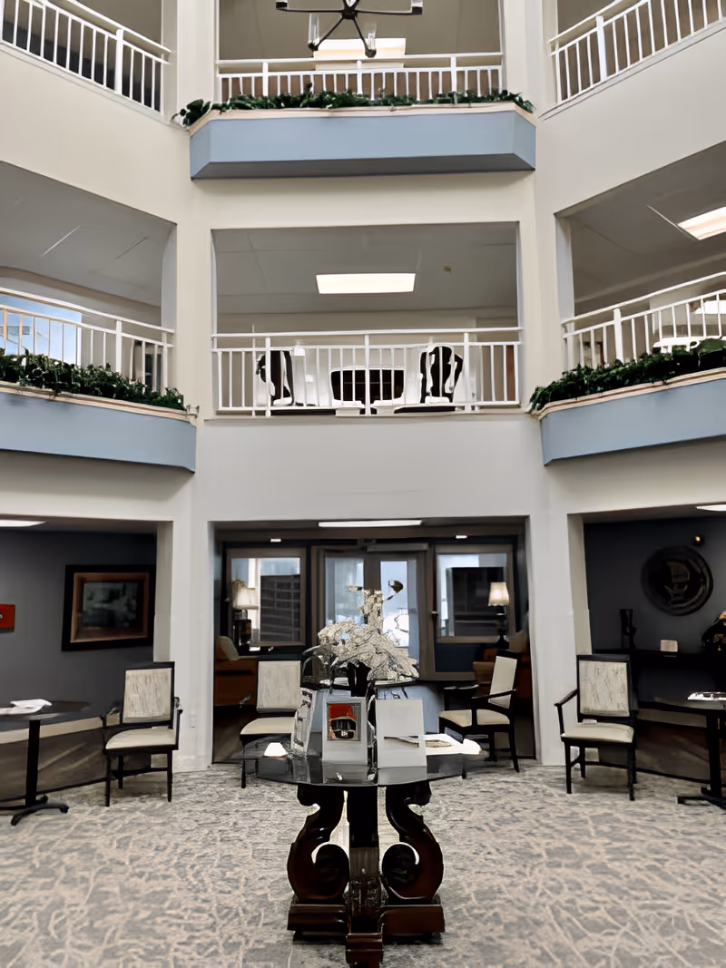 Interior view of a senior living facility lobby area with a central decorative table holding a silver sculpture and brochures. The space features multiple seating areas with chairs and small tables, a carpeted floor, and a two-story open atrium with white railings and greenery along the upper balcony edges.