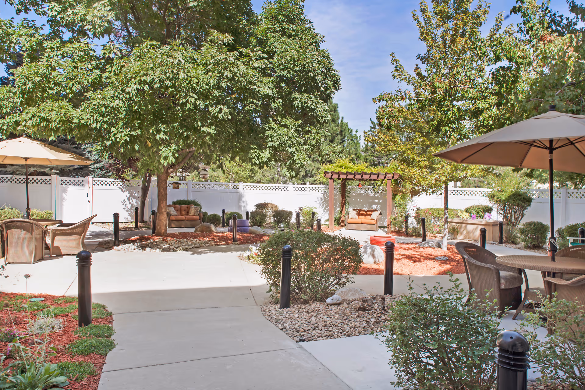 Courtyard with shaded seating areas, umbrellas, trees, and a pergola swing behind a white fence.