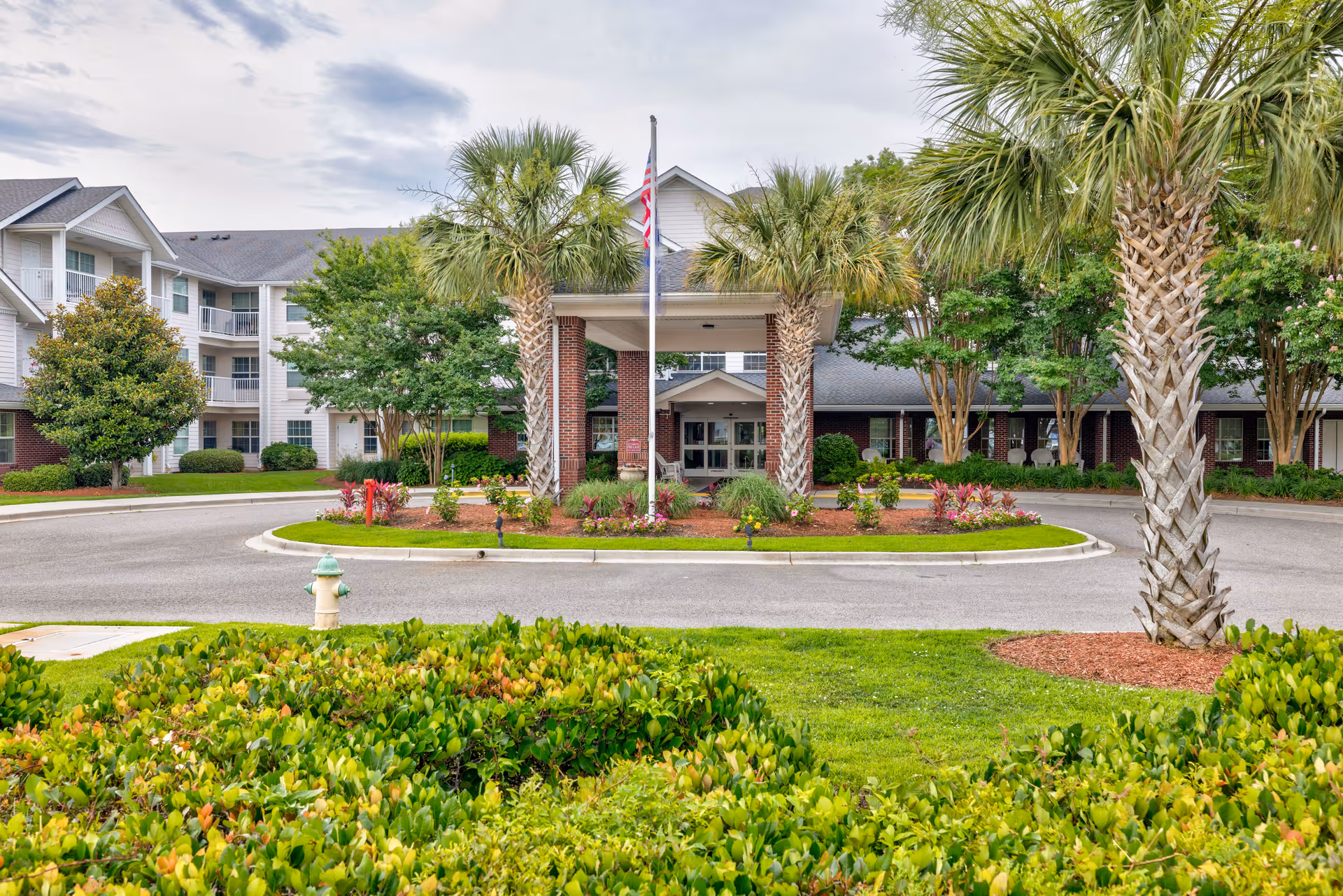Front exterior view of TerraBella Myrtle Beach facility with a circular driveway, landscaped greenery, palm trees, and an American flag in front of the entrance.