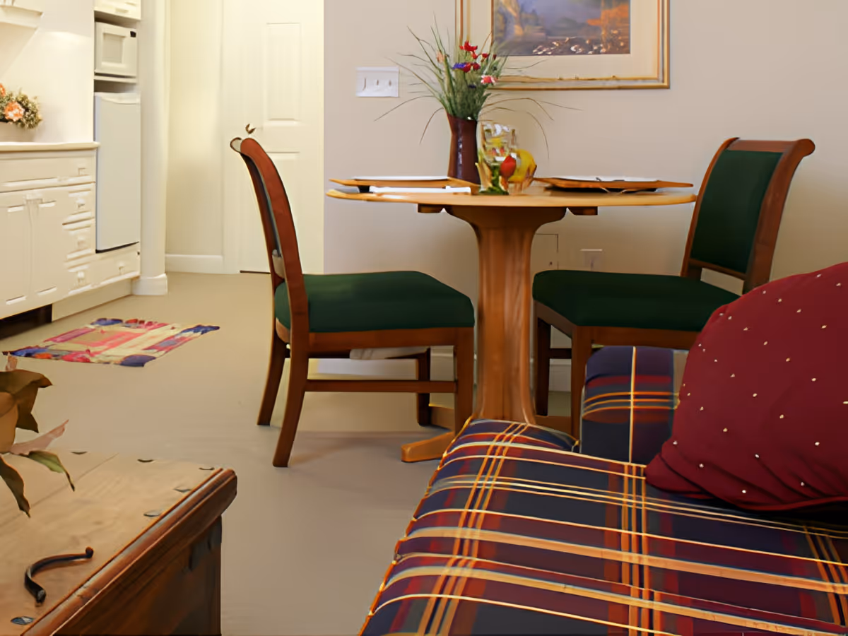 Small dining area with a round wooden table and green-upholstered chairs beside a kitchenette and a plaid sofa.
