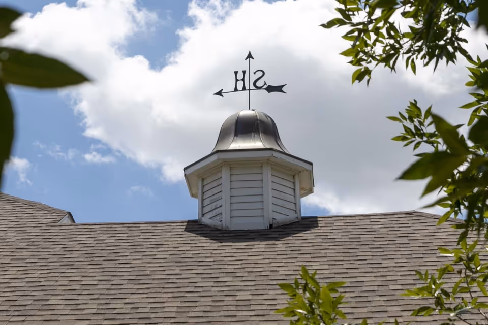Close-up view of a rooftop with a small cupola topped by a weather vane showing directions S and H, under a partly cloudy sky with some tree branches framing the image.