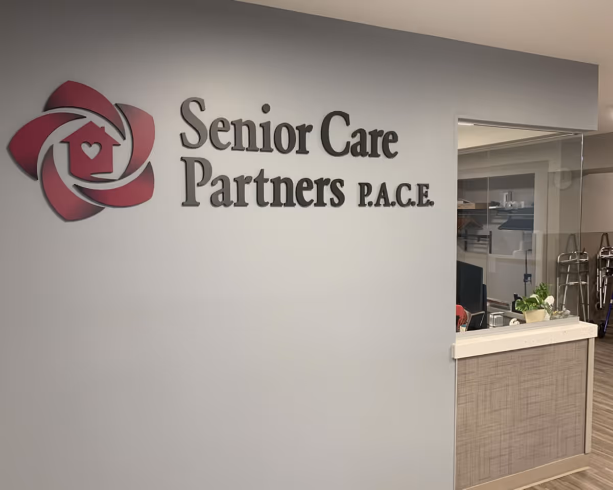 Interior reception area with a wall sign reading 'Senior Care Partners P.A.C.E.' and logo beside a small front desk.