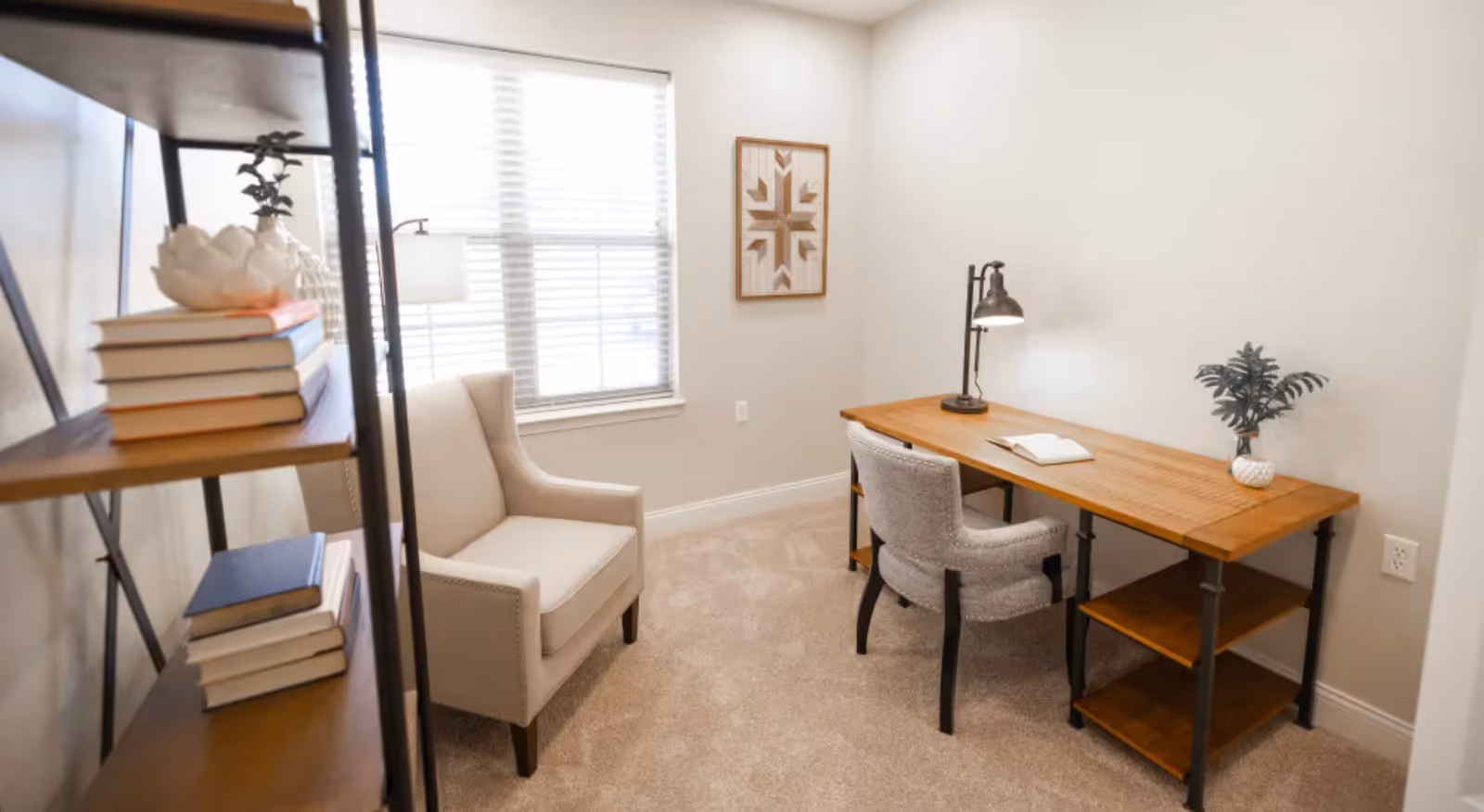 A small, cozy room with beige carpet and light-colored walls featuring a wooden desk with a lamp, an open book, and a small plant. There is a cushioned chair at the desk and a beige armchair near a window with blinds. A bookshelf with stacked books and decorative items is partially visible on the left side of the image. A framed geometric artwork hangs on the wall above the desk.