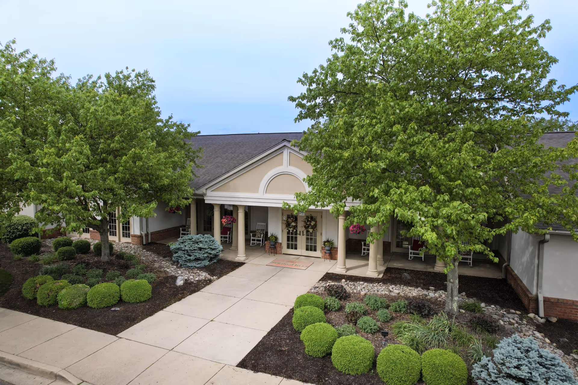 Front exterior view of a single-story building with a covered entrance supported by columns, surrounded by well-maintained landscaping including green bushes, trees, and flower pots near the entrance.