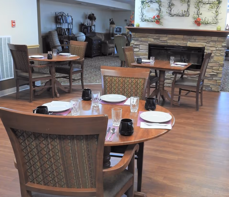 Dining room with wooden tables set for meals, chairs, and a stone fireplace in the background.
