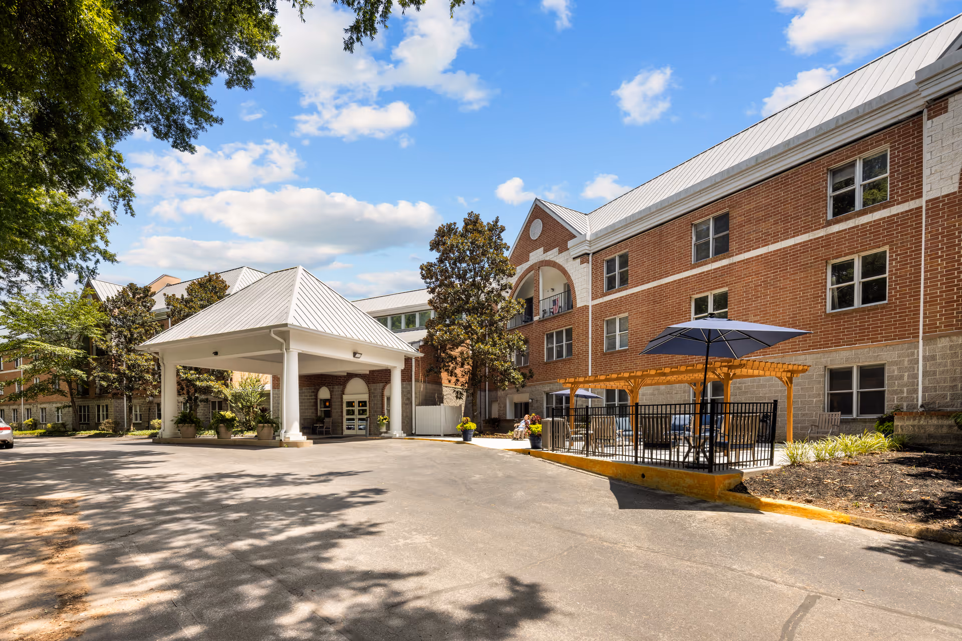 Front exterior of a brick senior living building with a covered entrance, driveway, and an outdoor seating area with umbrellas under a blue sky.