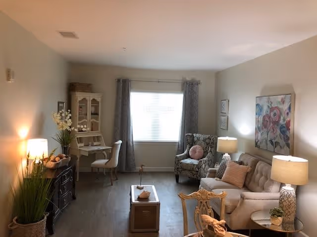 A cozy living room with a beige sofa adorned with pink and beige pillows, a floral armchair with a pink round pillow, two side tables each with a lamp, a coffee table in the center, a small desk with a chair and decorative items near the window with gray curtains, and a dark wooden cabinet with a lamp and plants on the left side.